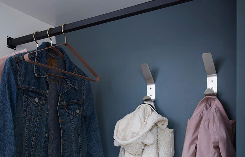 Mörkblå hatthylla med metallstång och krokar, upphängda: jeansjacka, fluffig vit jacka, rosa jacka.