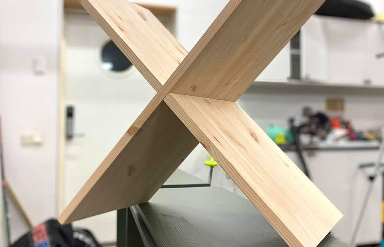 X-format korsade obehandlade ljusa träplankor på bänk i verkstad med verktyg och vit dörr