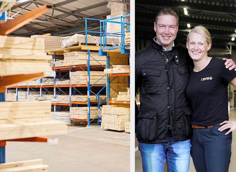 Lagerhall med staplade träreglar i blå-orange hyllor, två anställda i svarta jacka och t-shirt.