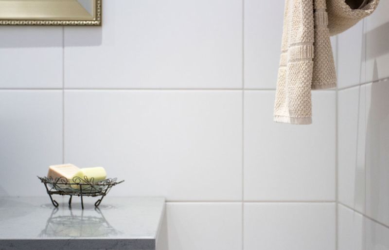 Vitt kakel, hängande beige handduk och tvålar i svart trådställ på ljus bänkskiva.