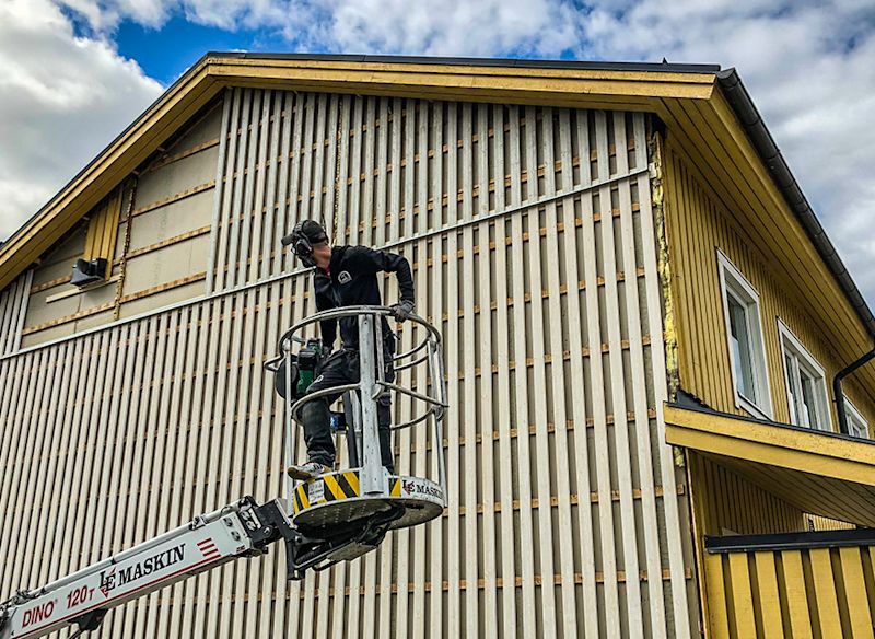 Arbetare på lift renoverar gul träfasad mot molnig grå himmel
