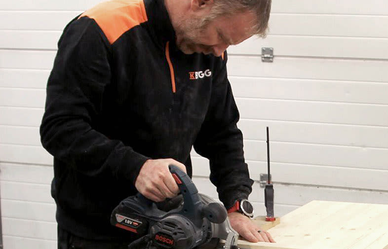 Person i svart-orange tröja kapar ljus träskiva med batteridriven Bosch-cirkelsåg i verkstad