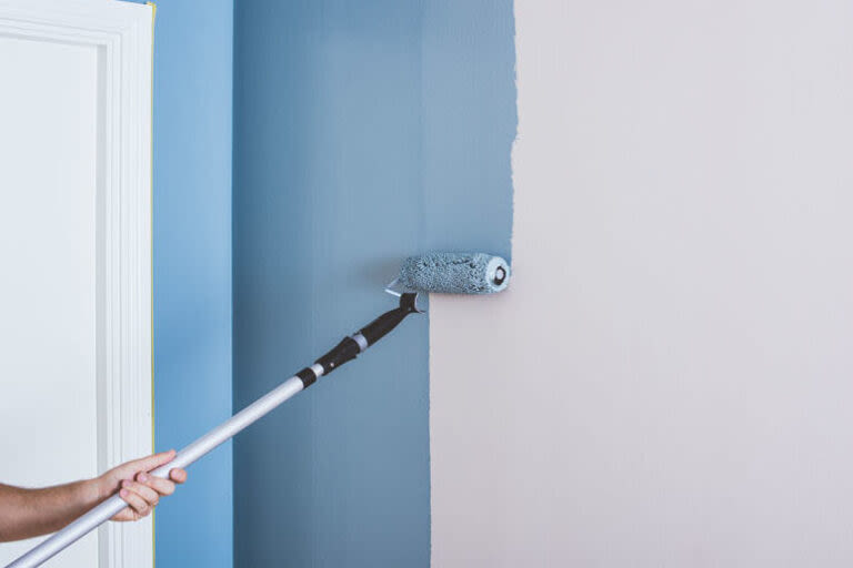 Arm med färgrulle på skaft målar inomhusvägg från vit till blå