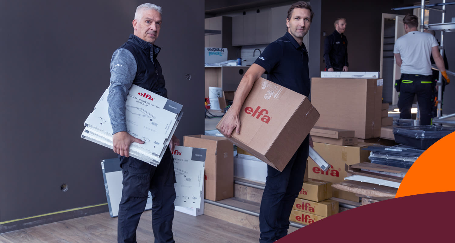 Två montörer bär Elfa-kartonger och installationsdelar i ett rum med mörka väggar.