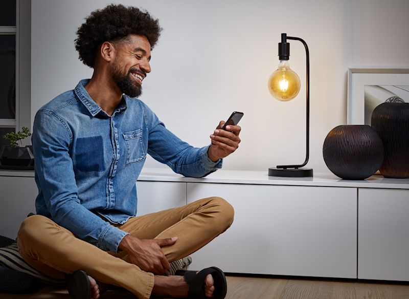 Man i jeansskjorta och beige byxor sitter med mobil vid vitt skåp, tänd lampa och svarta vaser.