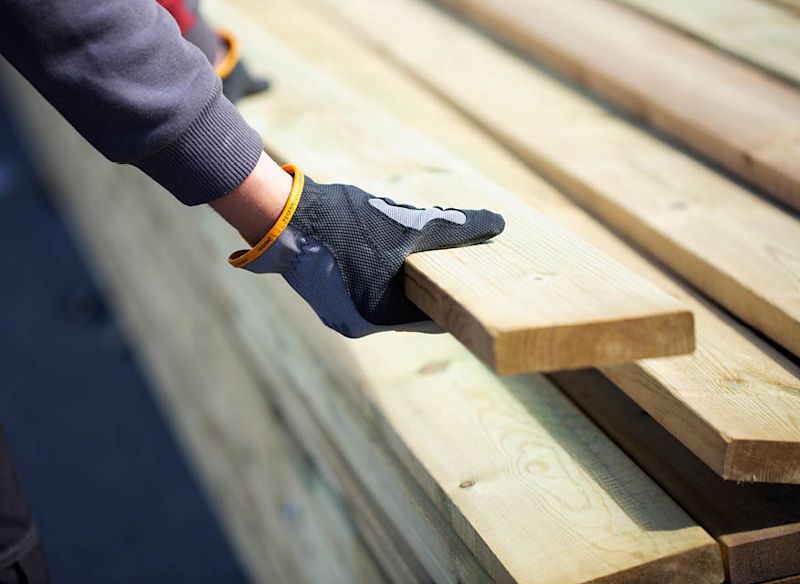 Svartgrå handske med gul kant lyfter en ljus träplanka från stapel av träbrädor.