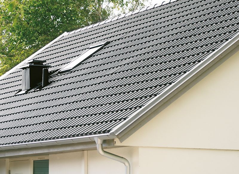 Ett hus med ett lutande tak av profilerad plåt. På taket finns ett takfönster och en svart skorsten/ventilationshuv. Fasaden är ljus och i förgrunden syns en hängränna. Bakgrunden består av träd med gröna blad.