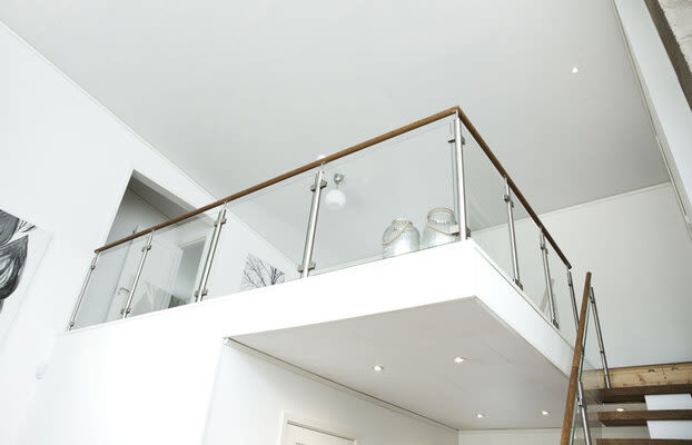 Vitt rum med loftbalkong i glasräcke och träledstång, trappa åt höger, minimalistisk stil.