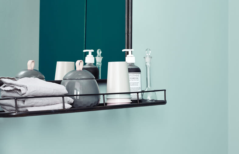 Svart hylla med grå handdukar, keramikkrukor, vit pumpflaska och klar glaskaraff mot turkos vägg