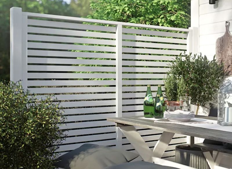 Vitt horisontellt spjälstaket bakom uteplats med träbord, gröna flaskor och växter.