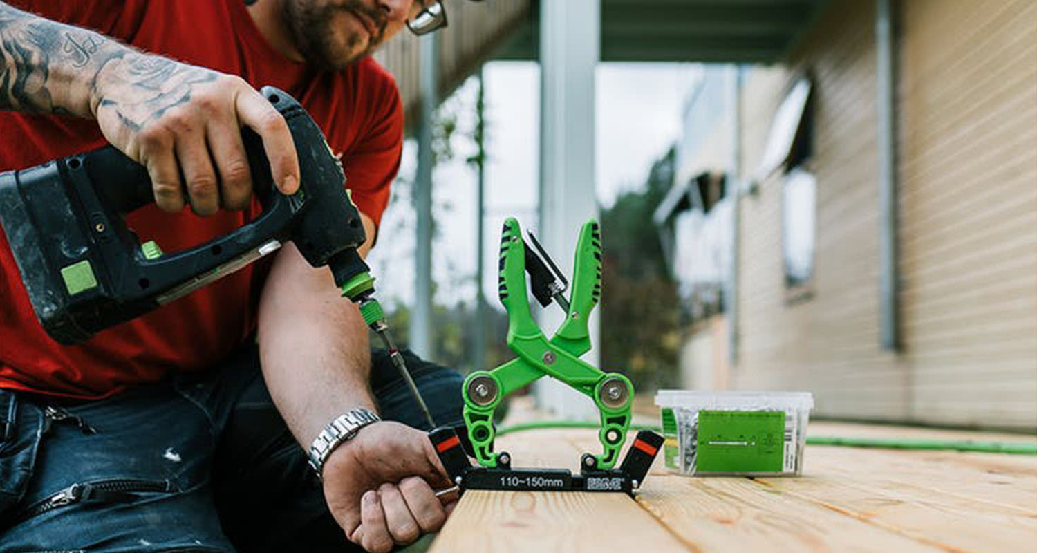 Byggarbetare i röd tröja borrar i trä med svart-grön borrmaskin och grön klämma.