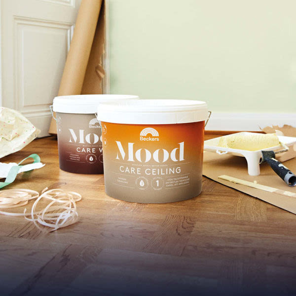 Två orange/bruna Mood Care Ceiling-färgburkar på parkettgolv med roller och tejp framför ljusgrön vägg.