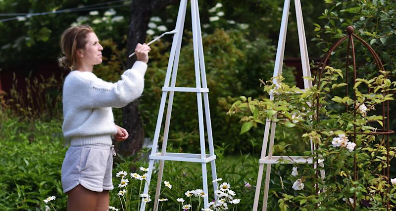 Person i vit tröja och grå shorts målar vit träobelisk bland prästkragar i frodig trädgård.