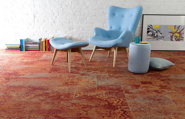Ljusblå fåtölj med fotpall och liten pall på rödorange mönstrad matta framför vit tegelvägg.