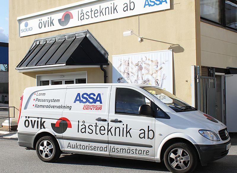 En vit servicebil med texten "Övik låsteknik ab" står parkerad framför en byggnad med samma företagsnamn på skylten. Text på bilen nämner larm, passersystem och kameraövervakning.