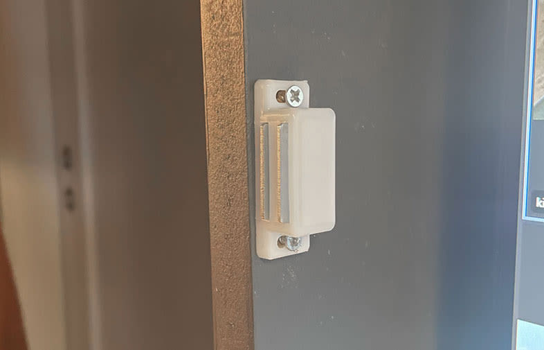 Vit plastdörrsensor med skruvar monterad på mörkgrå dörrstomme intill träfoder.