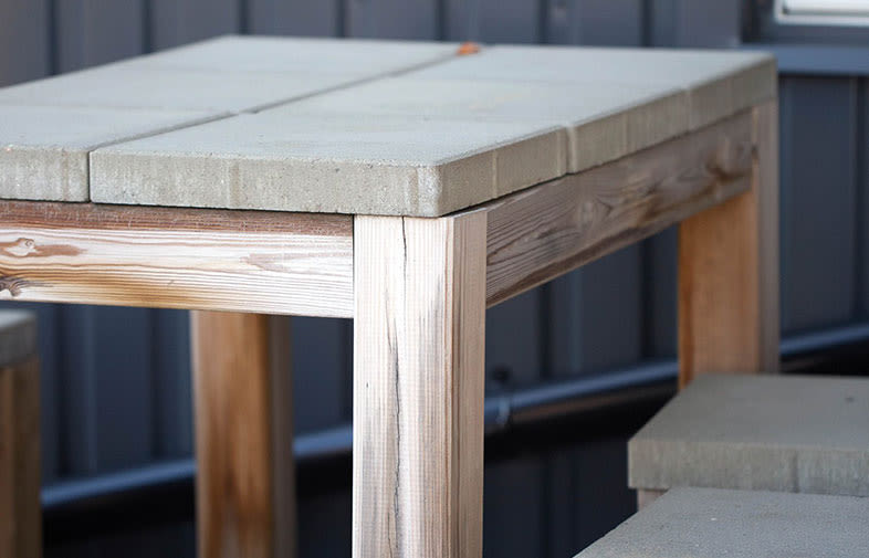 Högt utebord i ljust trä med grå betongskivor mot mörkgrå vägg