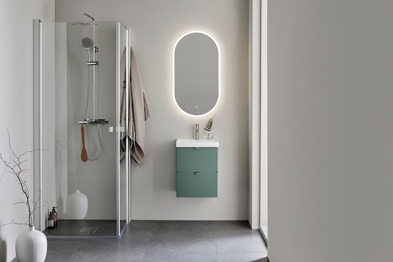 Modernt grått badrum med glasdusch, hängande handduk, grönt skåp under oval spegel med belysning.