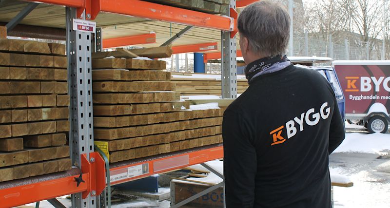 Man i svart K-BYGG-tröja granskar staplade träbalkar på orange hyllor utomhus i snö