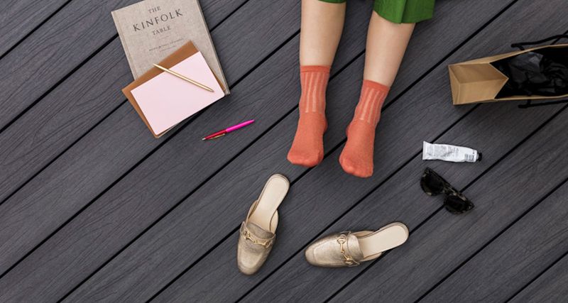 Ovanifrån: fötter i orange strumpor på mörkt trädäck omgivna av guldsandaler, bok och papperspåse