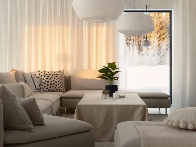 Vardagsrum med beige hörnsoffa, vita hänglampor, växt på dukat bord, och snötäckt skog utanför.