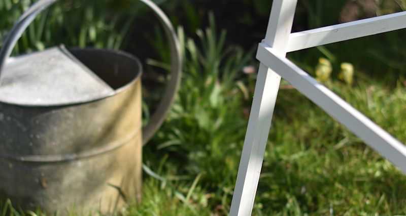 Grå metallvattenkanna vid vitt trädgårdsstativ på grön gräsmatta.