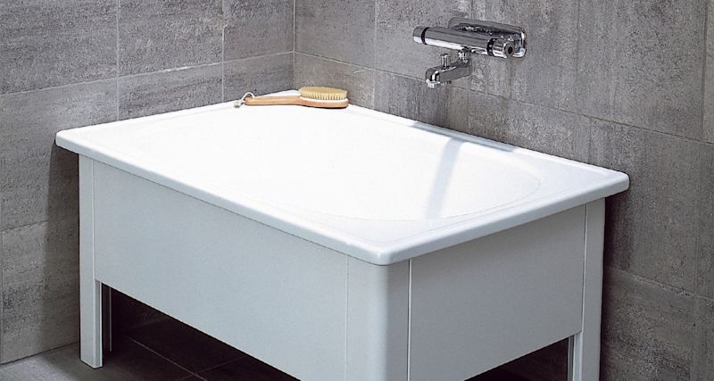 Vit rektangulär tvättbalja med träborste på kanten, grå kakelvägg och kromad väggblandare.