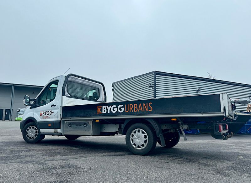 Vit flakbil med svart flak märkt 'KBYGG Urbans' står på asfalt framför industribyggnad under grå himmel