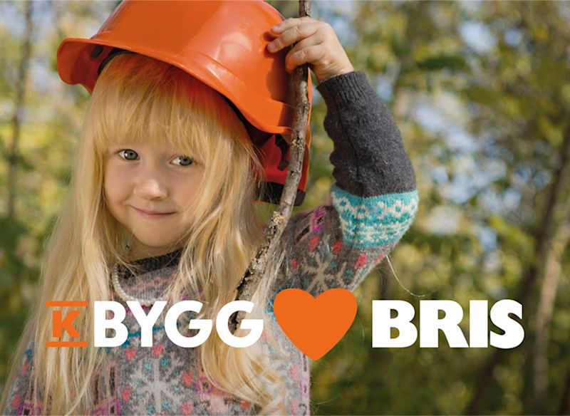 Ett barn står utomhus i en skogslik miljö, bär en orange bygghjälm och håller i en pinne. I förgrunden syns texten “K-Bygg ❤️ Bris”.