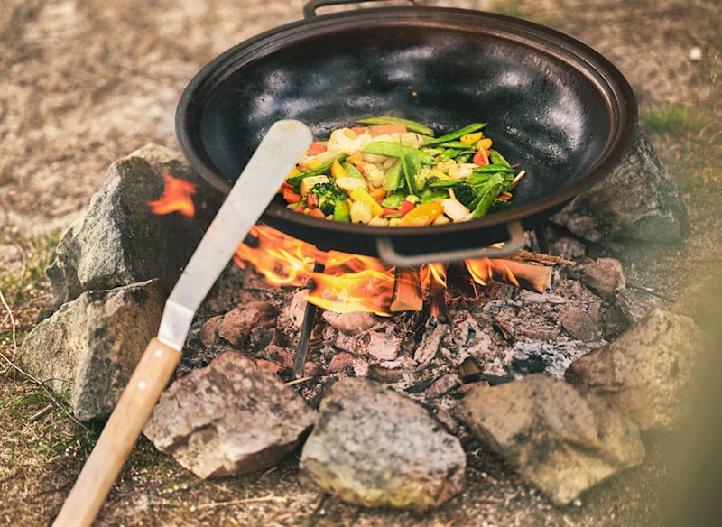Svart wokpanna över lägereld omgiven av stenar, med färgglada grönsaker och spatel.