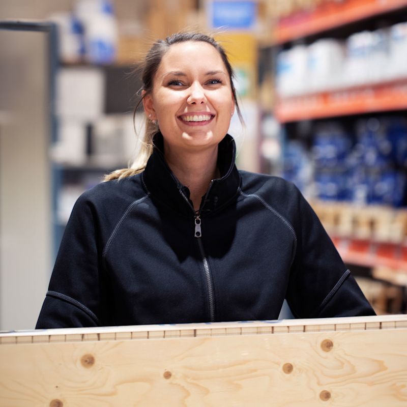 Person i svart jacka håller träskiva i byggvaruhandel med färg- och verktygshyllor.