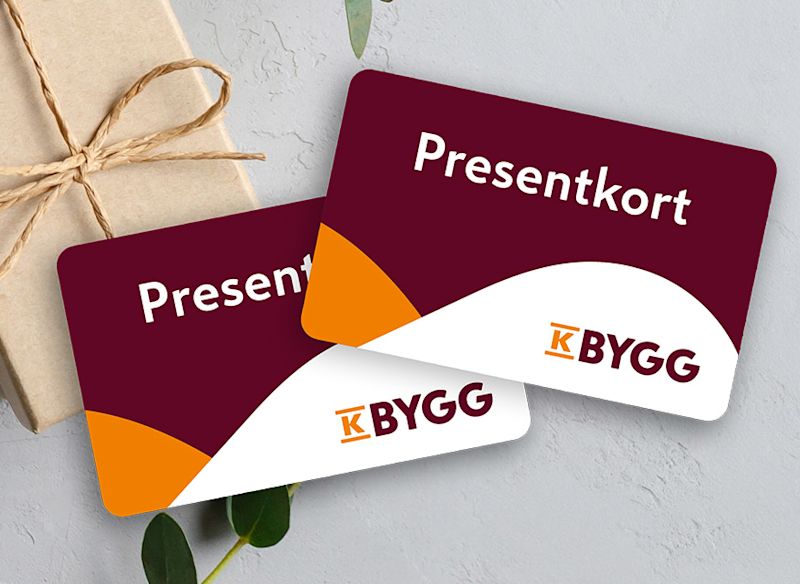 Två K-Bygg-presentkort i vinrött, vitt och orange på ljusgrå bakgrund bredvid inslaget paket