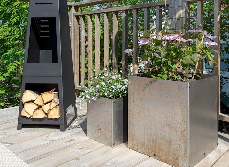 Svart metallkamin med ved, två rostiga grå lådor med vita och rosa blommor på trädäck.