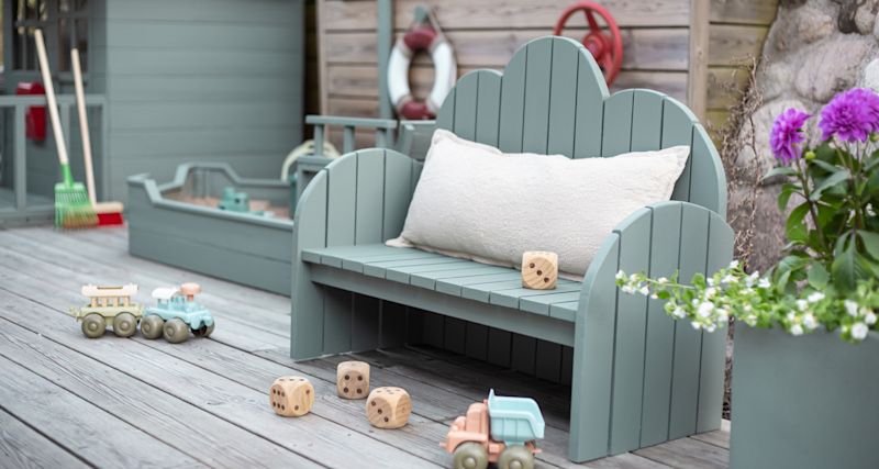 Mintgrön träbänk med vit kudde, trätärningar, leksakståg och lila blommor på trädäck.