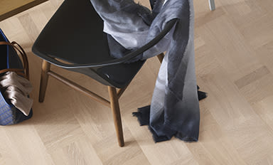 Svart stol med träben, grå sjal slängd över, blå väska med beige tyg på parkettgolv.