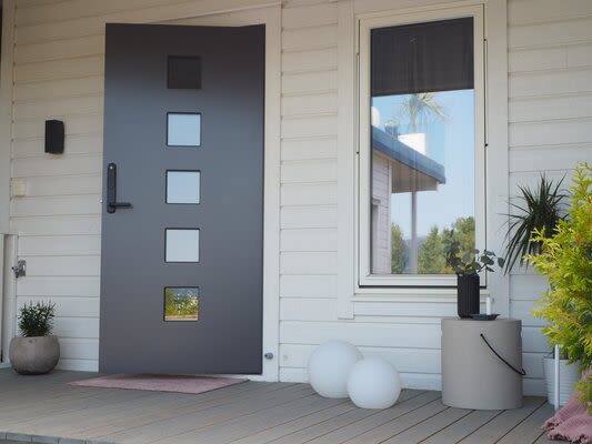 Modern mörkgrå dörr med fyra fyrkantiga fönster i vit träfasad, krukväxter och vita bollar.