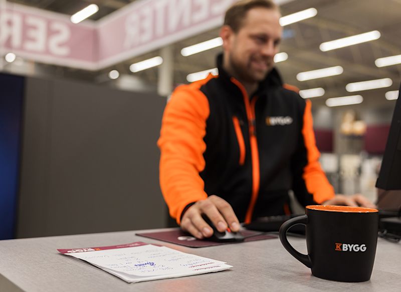 Medarbetare på K-Bygg arbetar vid kassan med dator och mus. På disken ligger anteckningar och en kaffemugg med K-Byggs logotyp, i en modern butiksmiljö.