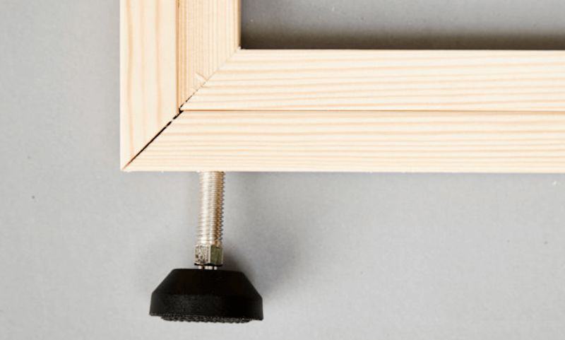 Hörn av ljus träram med silverfärgad gängstång och svart justerbar fot mot grå bakgrund