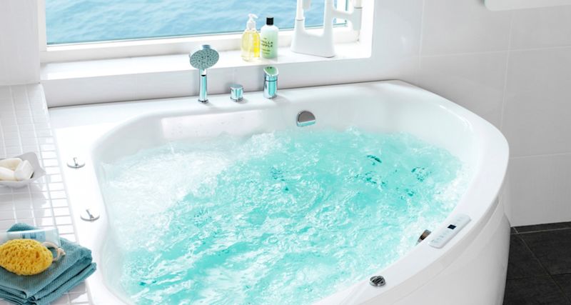 Vitt hörnbubbelbad med turkost porlande vatten vid fönster, blå handdukar och tvål.