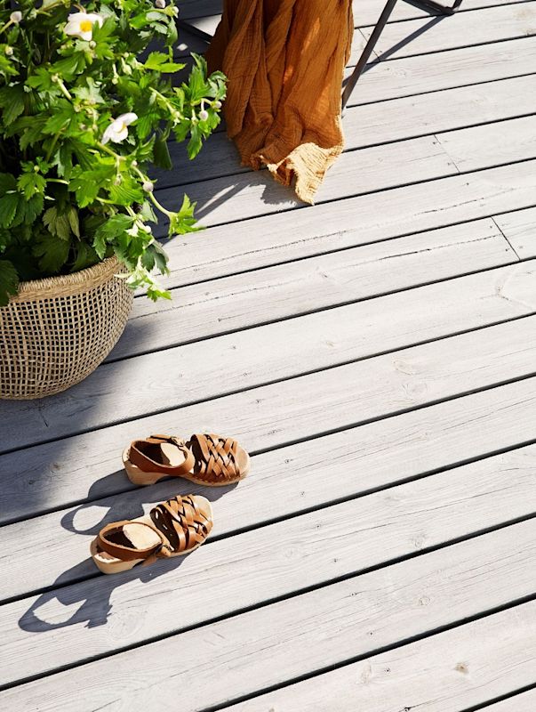 Bruna flätade sandaler på ljus trätrall vid kruka med gröna blad, vita blommor och orange filt