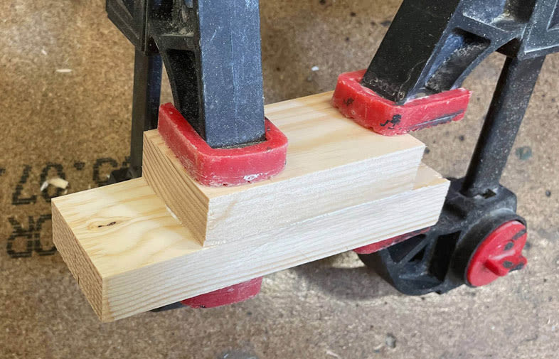 Svart plastskruvtving med röda glidskydd fäster två ljusa träbitar på en snickarbänk.