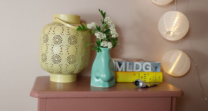 Rosa bord med gul perforerad lykta, grön näsvas med vita blommor, böcker och ljusslinga