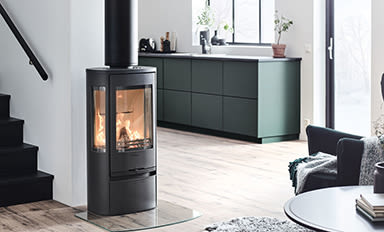 Svart glasinsatskamin med sprakande eld i ljust vardagsrum med gröna köksskåp