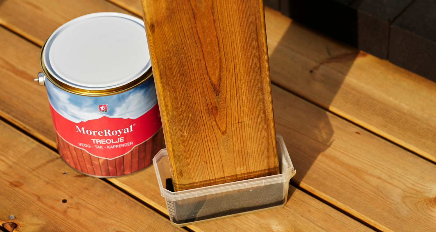 Röd-blå burk MoreRoyal träolja och träplanka doppad i olja på solbelyst trädäck.