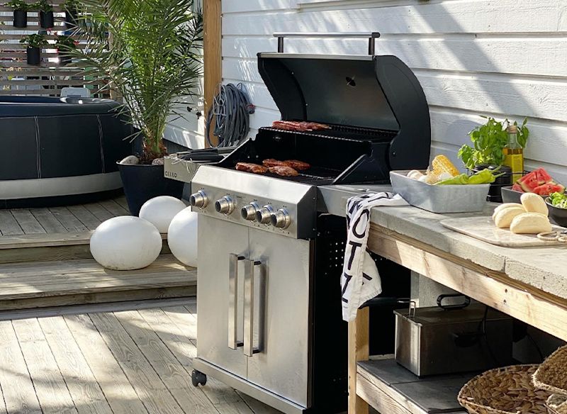 Svart gasolgrill med kött på trädäck, intill bord med färska grönsaker, bröd och olivolja.