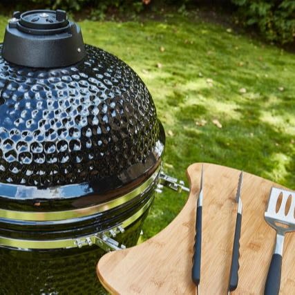 Närbild på svart keramisk grill med trähylla och grillredskap på grön gräsmatta