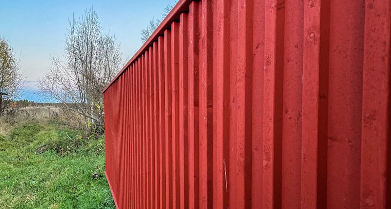 Röd korrugerad plåtvägg vid grönt gräs och kala buskar under klarblå himmel