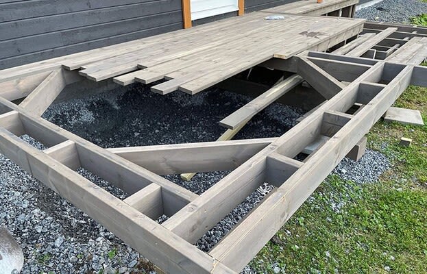 Delvis monterad grå träram för altan med trallbrädor och grus under intill svart fasad.