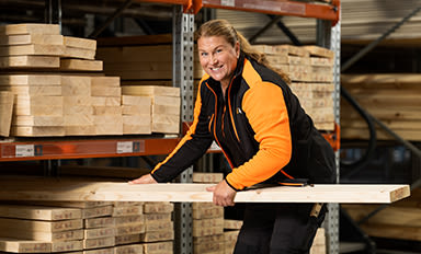 Person i orange-svart arbetsjacka hanterar träplanka i trävarulager