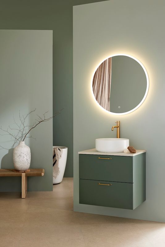 Badrum med rund varmvitt belyst spegel, vit skålhandfat, guldfärgad blandare och mörkgrön kommod.